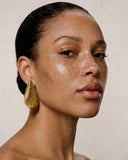 A close-up portrait of a woman with dark hair pulled back, wearing a large textured gold teardrop earring, against a warm beige background.