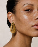 A close-up portrait of a woman with dark hair pulled back, wearing a large textured gold teardrop earring, against a warm beige background.